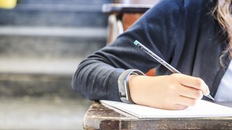 Eine Person schreibt mit einem Stift auf einen Zettel, im Hörsaal sitzend